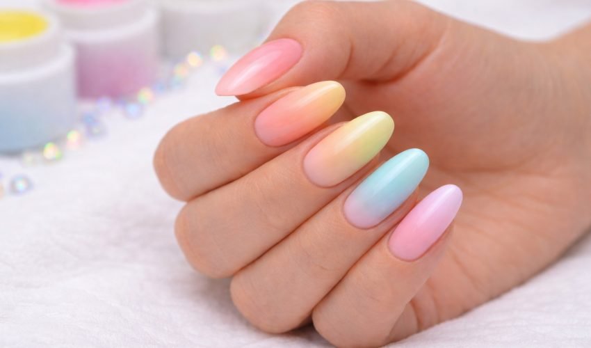 Main féminine avec vernis semi-permanent en dégradé pastel coloré, style baby-color, sur fond neutre beige.